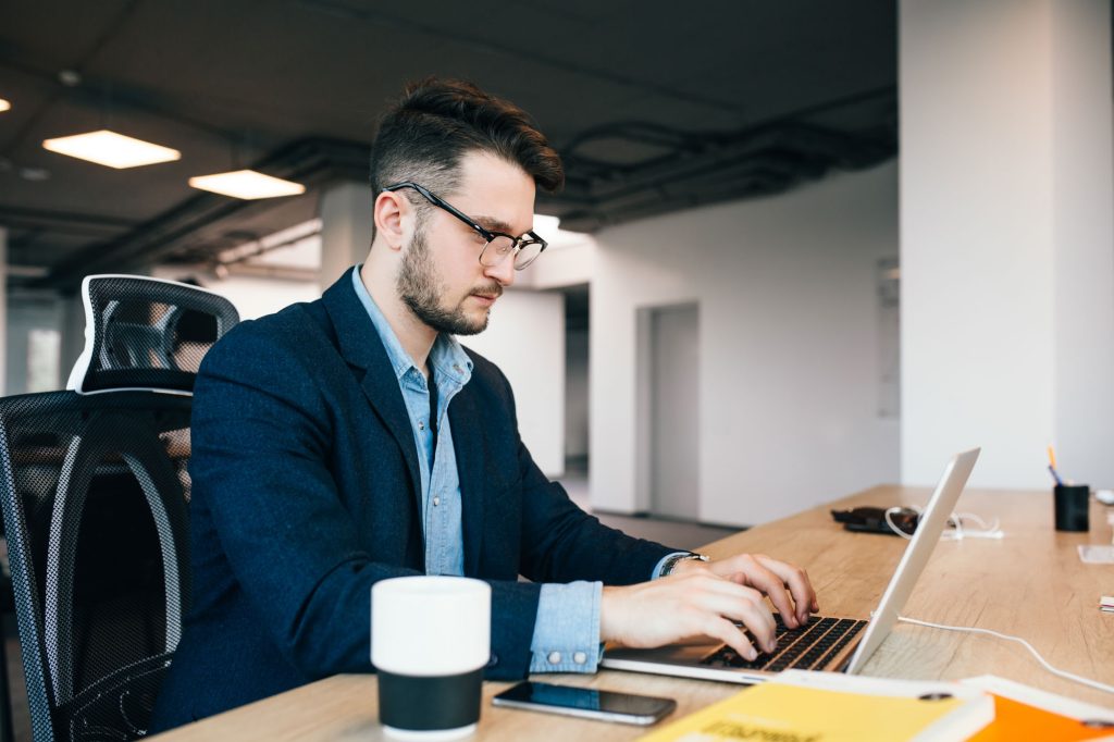 young dark haired man is working table office he wears blue shirt with black jacket he is typing laptop looks busy